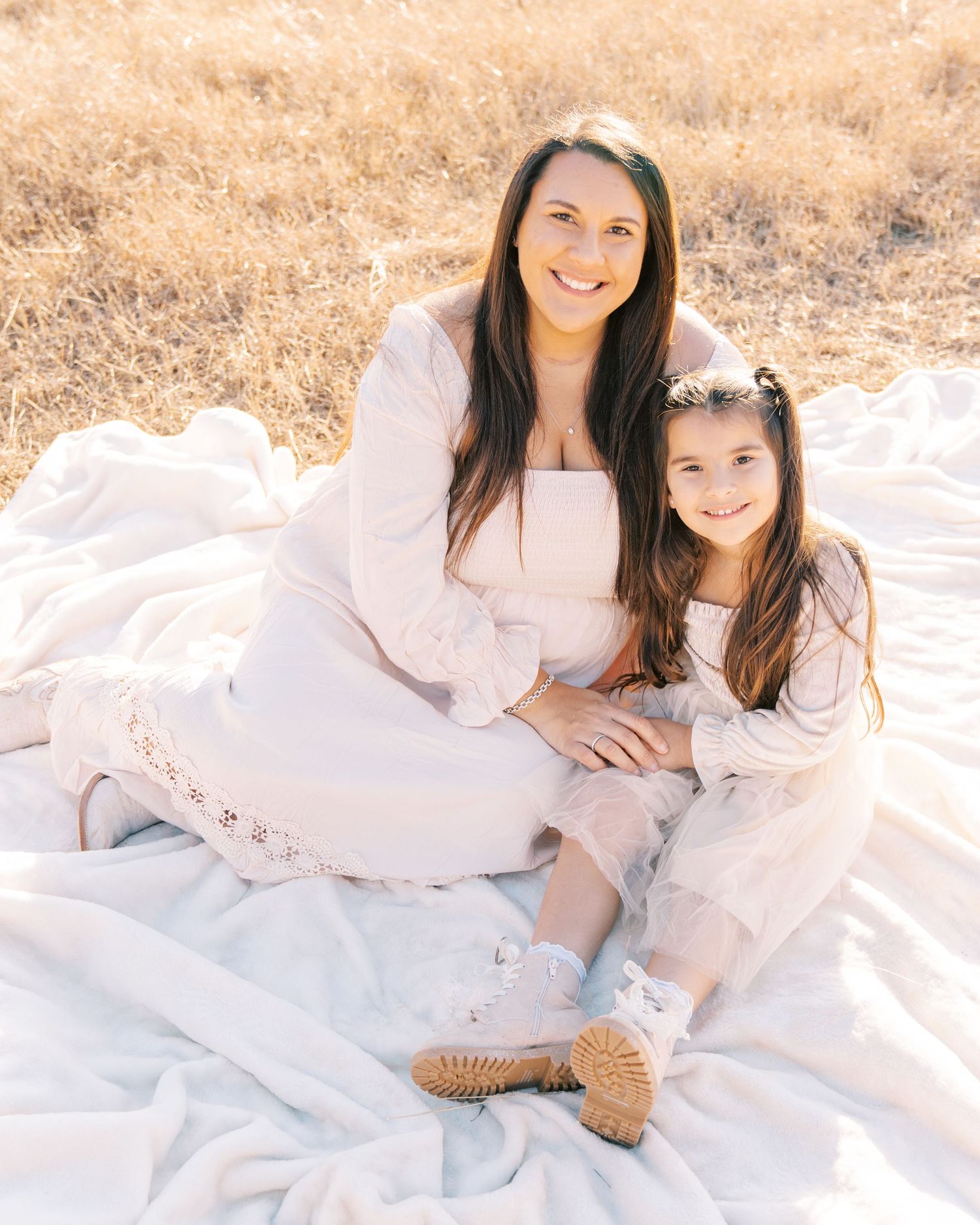 Two people in white dresses sitting together on a blanket in a sunny outdoor setting.