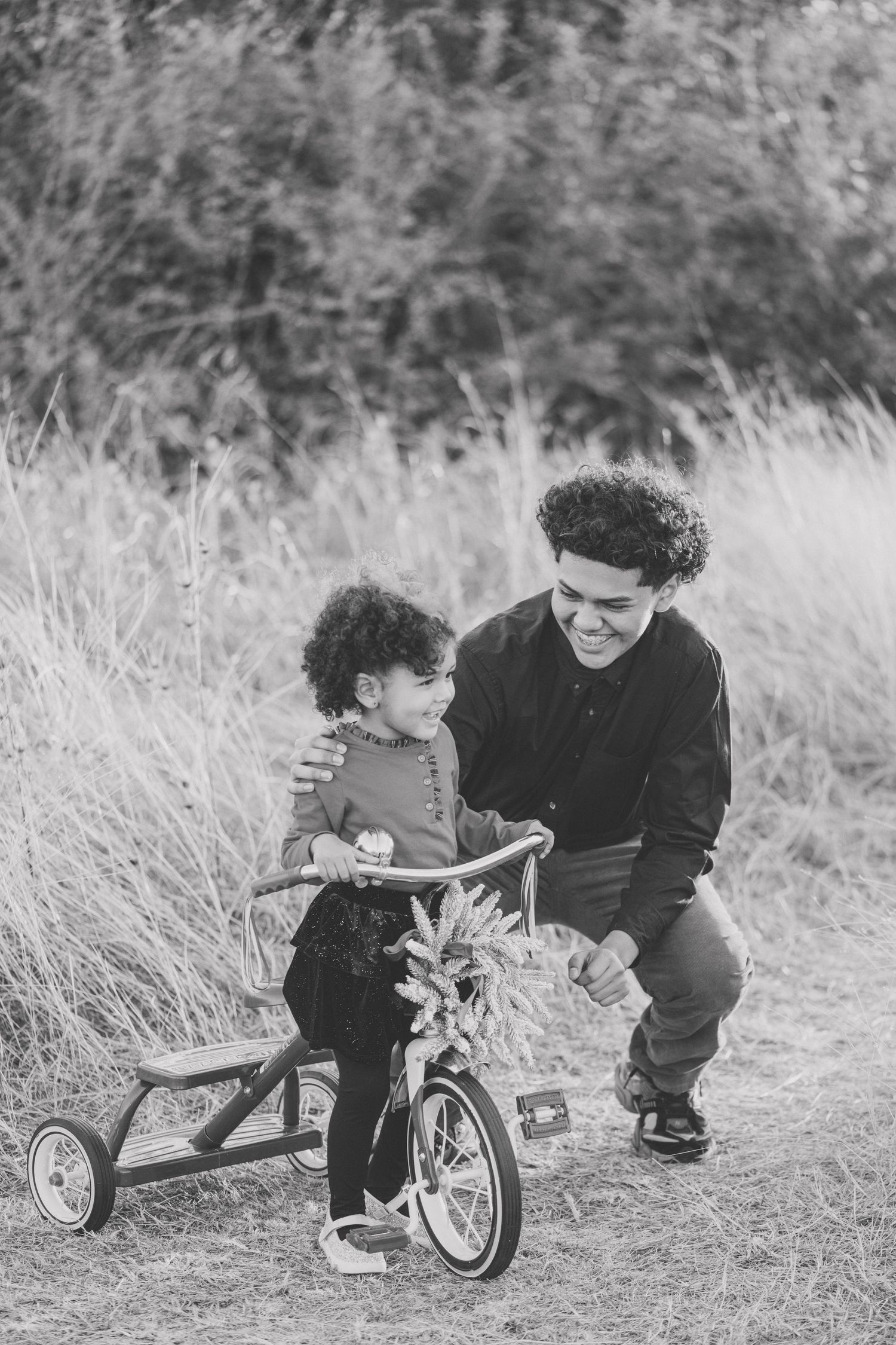 Black and white photo of small child learning to ride tricycle with adult guidance in field.