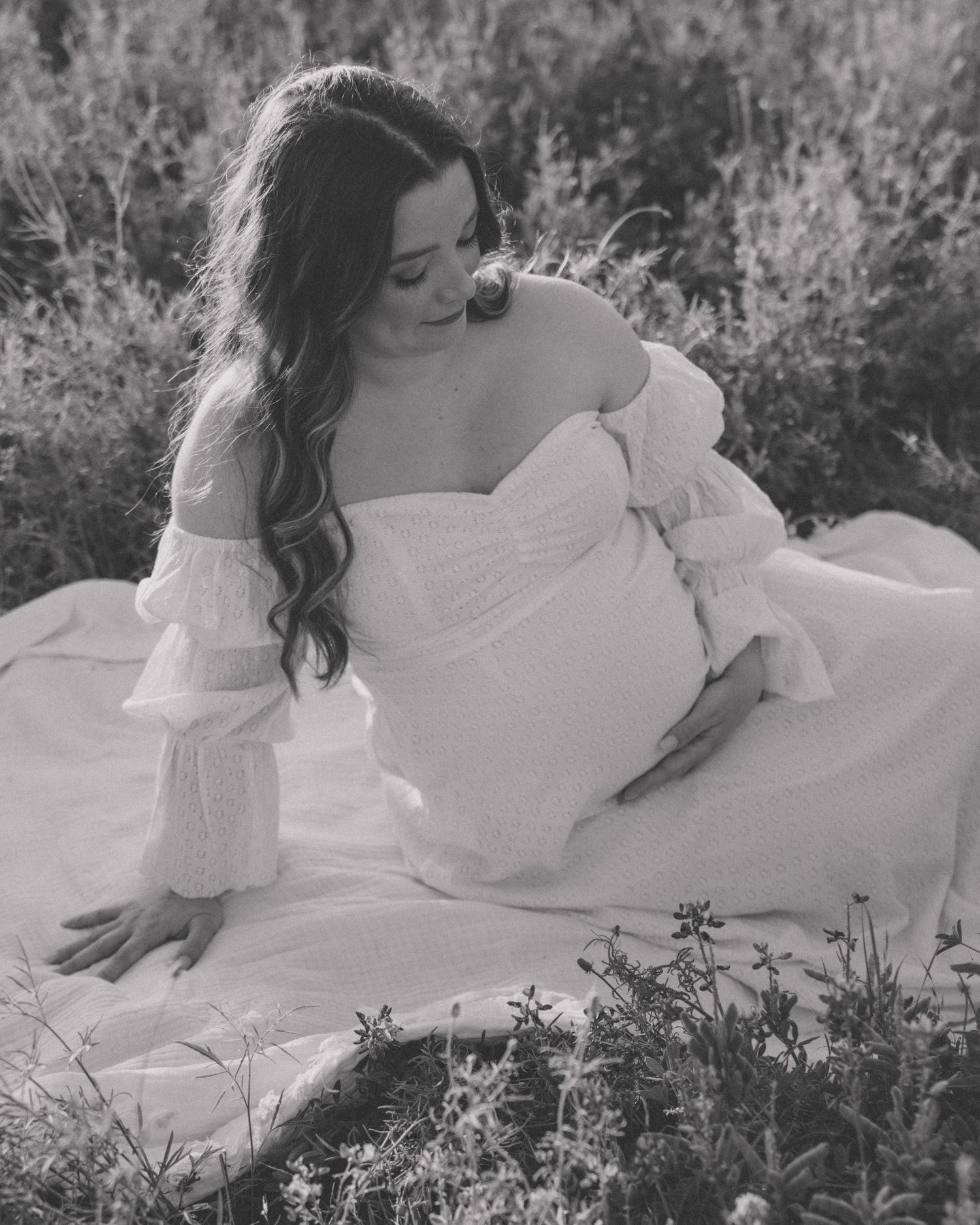 Black and white maternity photo of a woman in a white off-shoulder dress sitting in a wildflower field.