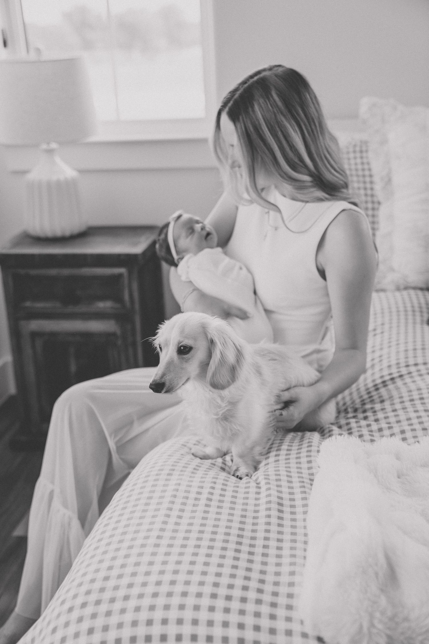 Black and white photo of a woman holding a newborn baby on a bed with a fluffy dachshund dog nearby.