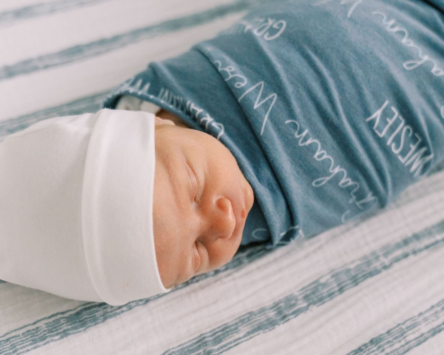 Newborn baby swaddled in white wrap wearing a white hat, resting on a personalized blue striped blanket.