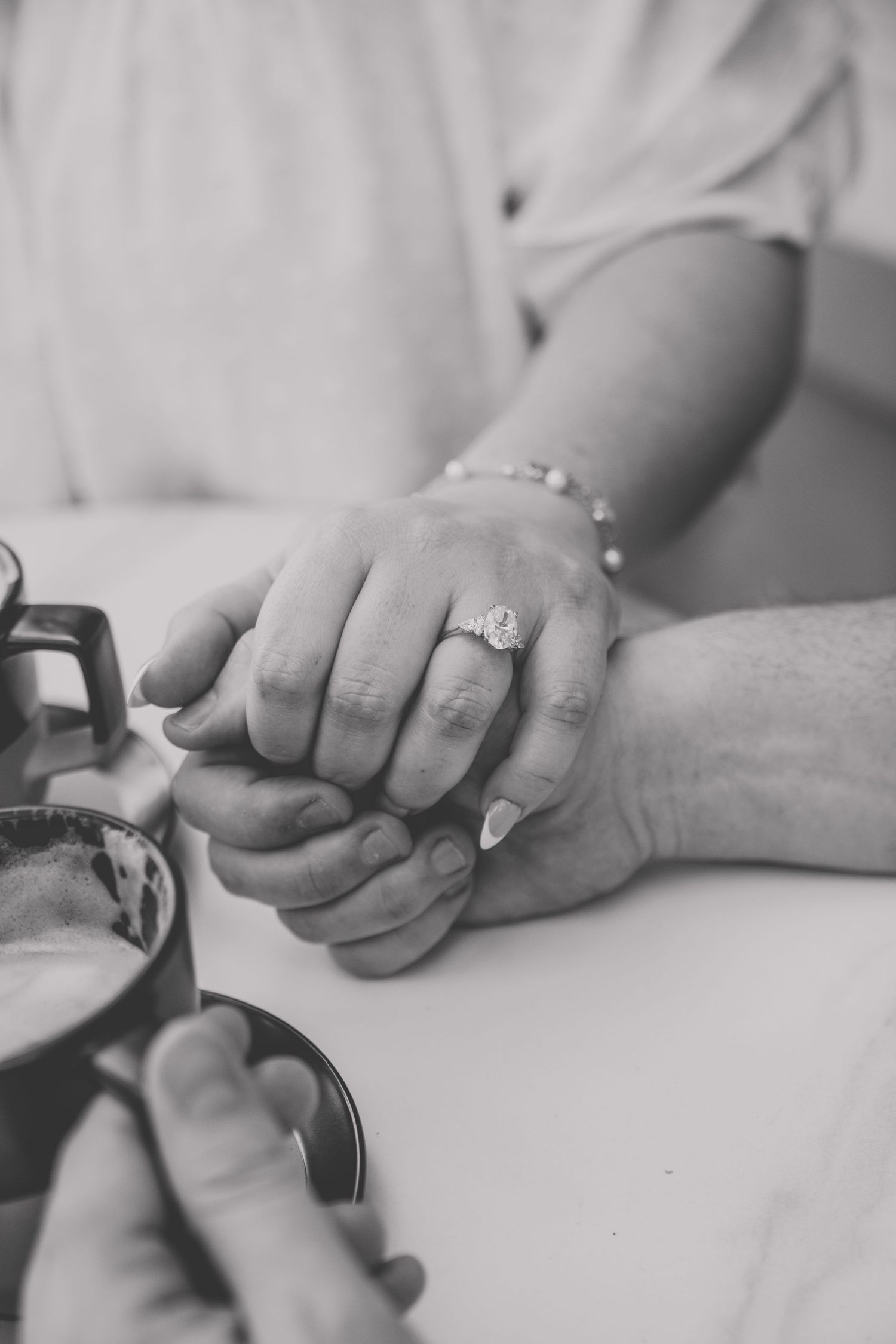 Black and white photo of couple holding hands, woman wearing diamond engagement ring and pearl bracelet at table.