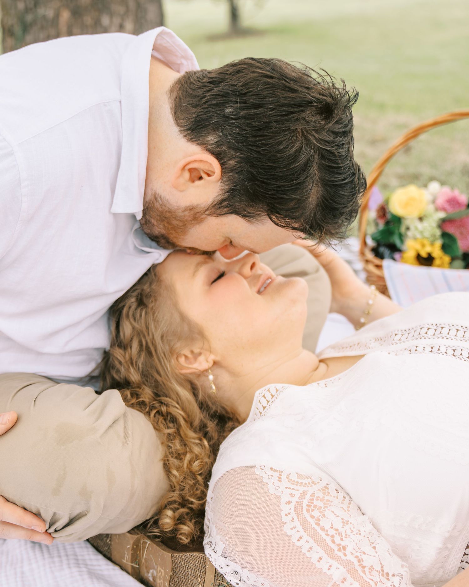 Man in white shirt leaning down to kiss woman on forehead during romantic outdoor picnic engagement session.