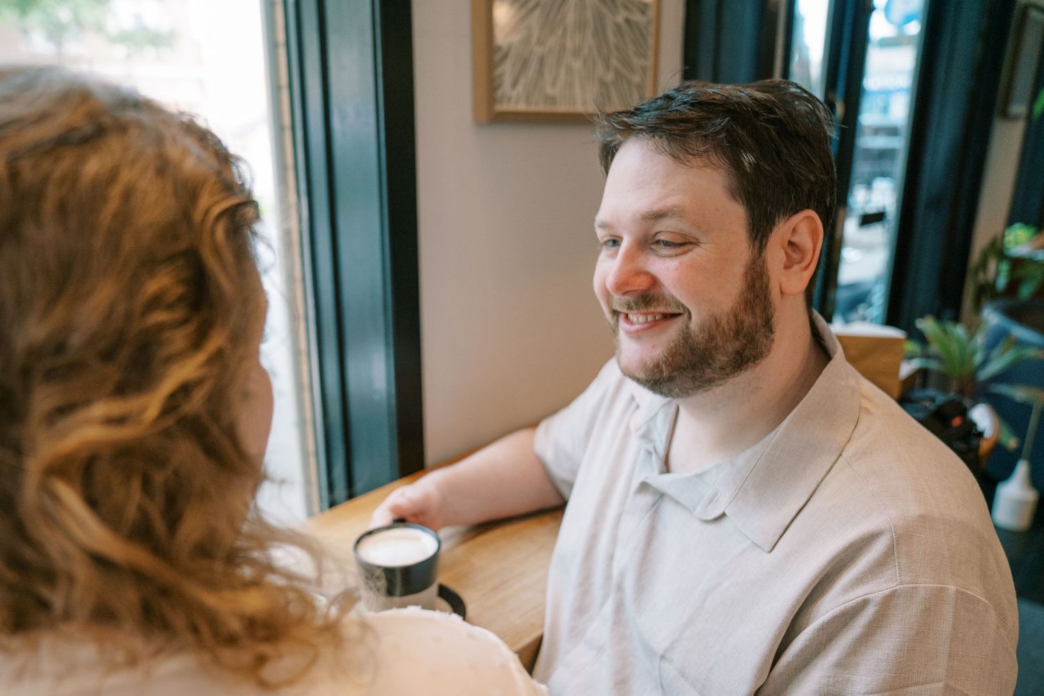 Smiling bearded man in beige polo shirt enjoying coffee at a cafe table during a couples date.