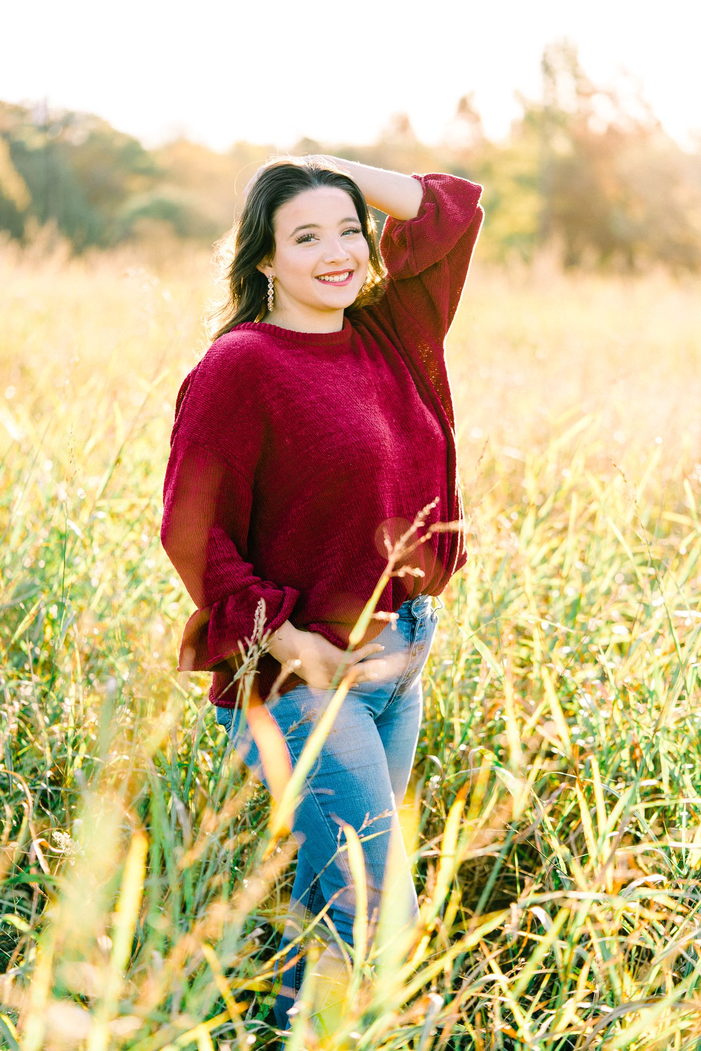 Person wearing burgundy sweater and jeans poses joyfully in a field during golden hour.