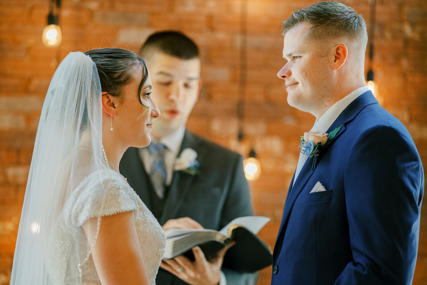 A couple exchanges vows during a wedding ceremony inside a rustic brick-walled venue with warm lighting.