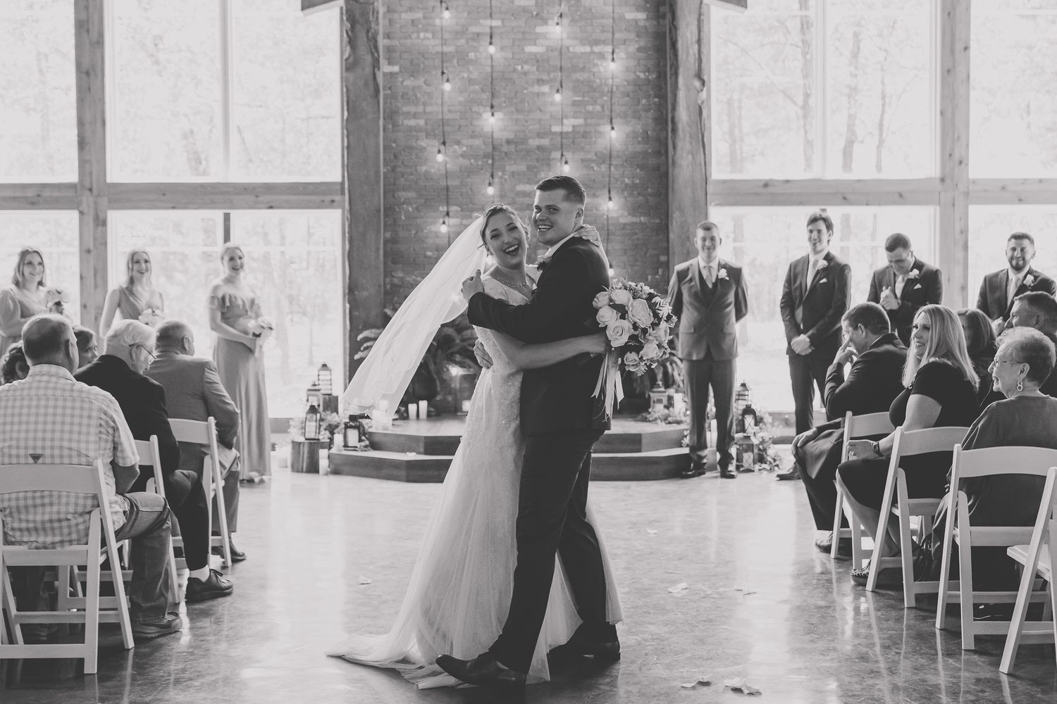A wedding ceremony takes place in an elegant ballroom with white chairs and twinkling lights overhead.