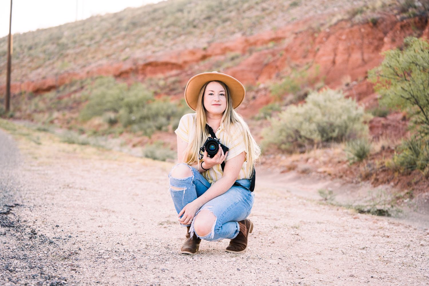 A person in a straw hat and denim poses on a winding dirt path with red rock formations in the background.