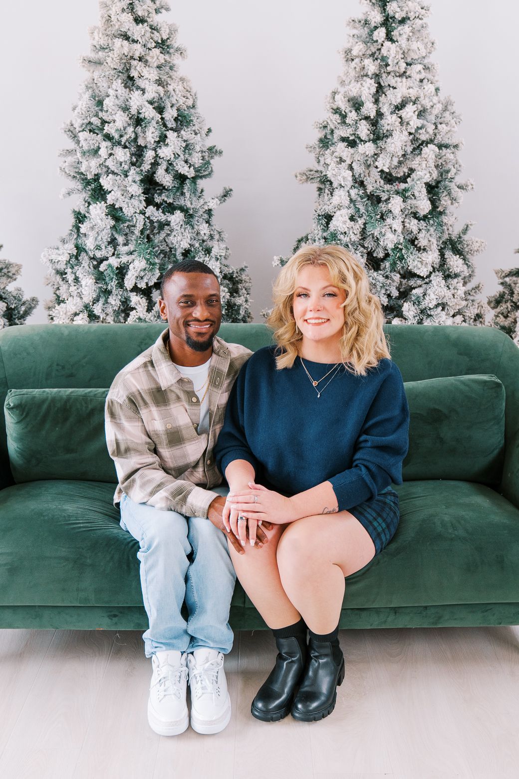 A couple sits together on a green velvet couch with decorated Christmas trees in the background.