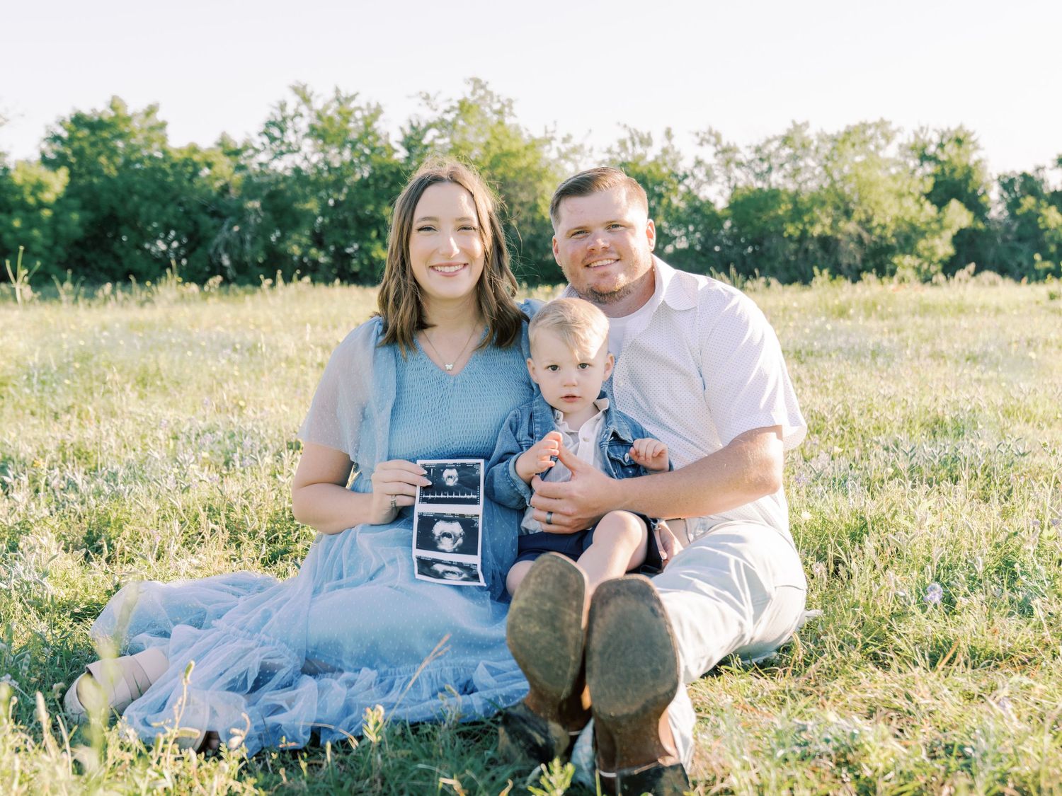 A family sits together in a sunlit meadow holding ultrasound photos during a pregnancy announcement photoshoot.