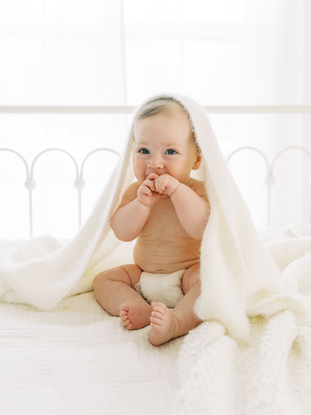 Baby wrapped in white blanket playing peek-a-boo on bright white bedding.