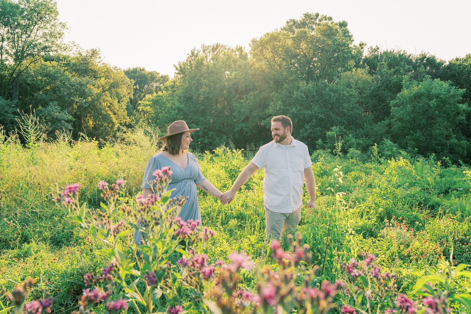 A couple walks hand in hand through a vibrant meadow filled with wildflowers and greenery at sunset.