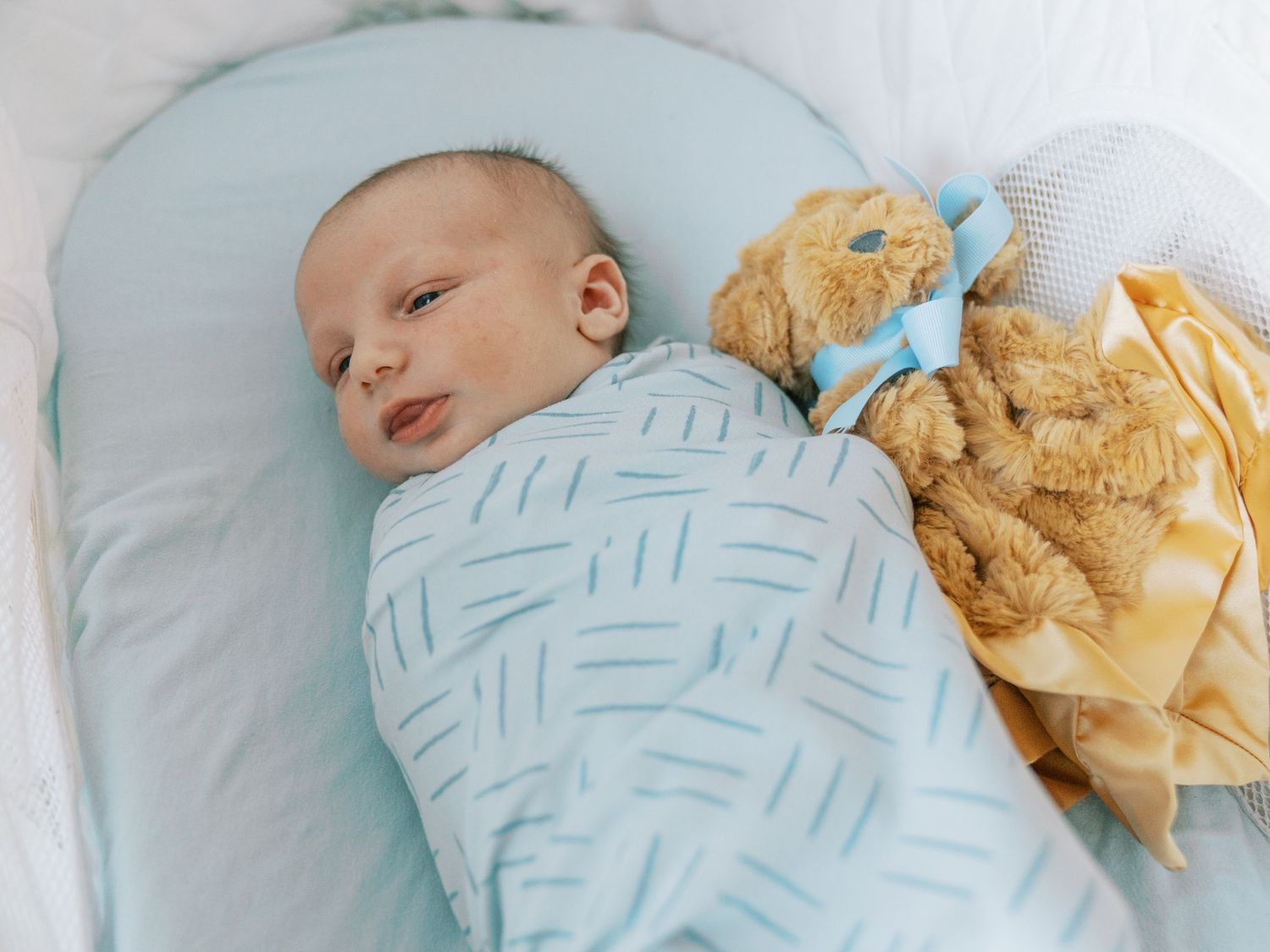Baby in light blue swaddle rests next to a plush toy on white bedding.
