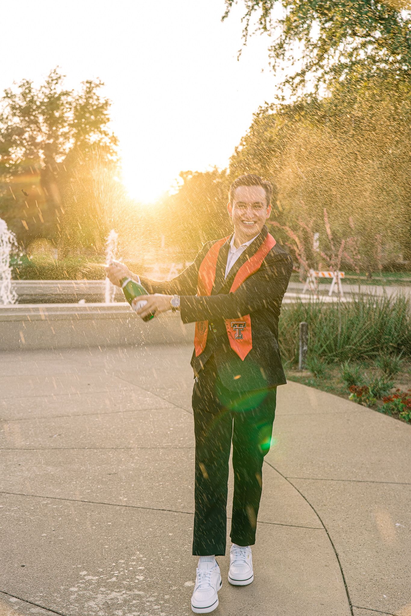 Graduate in cap and gown pops champagne during golden sunset celebration on campus pathway.
