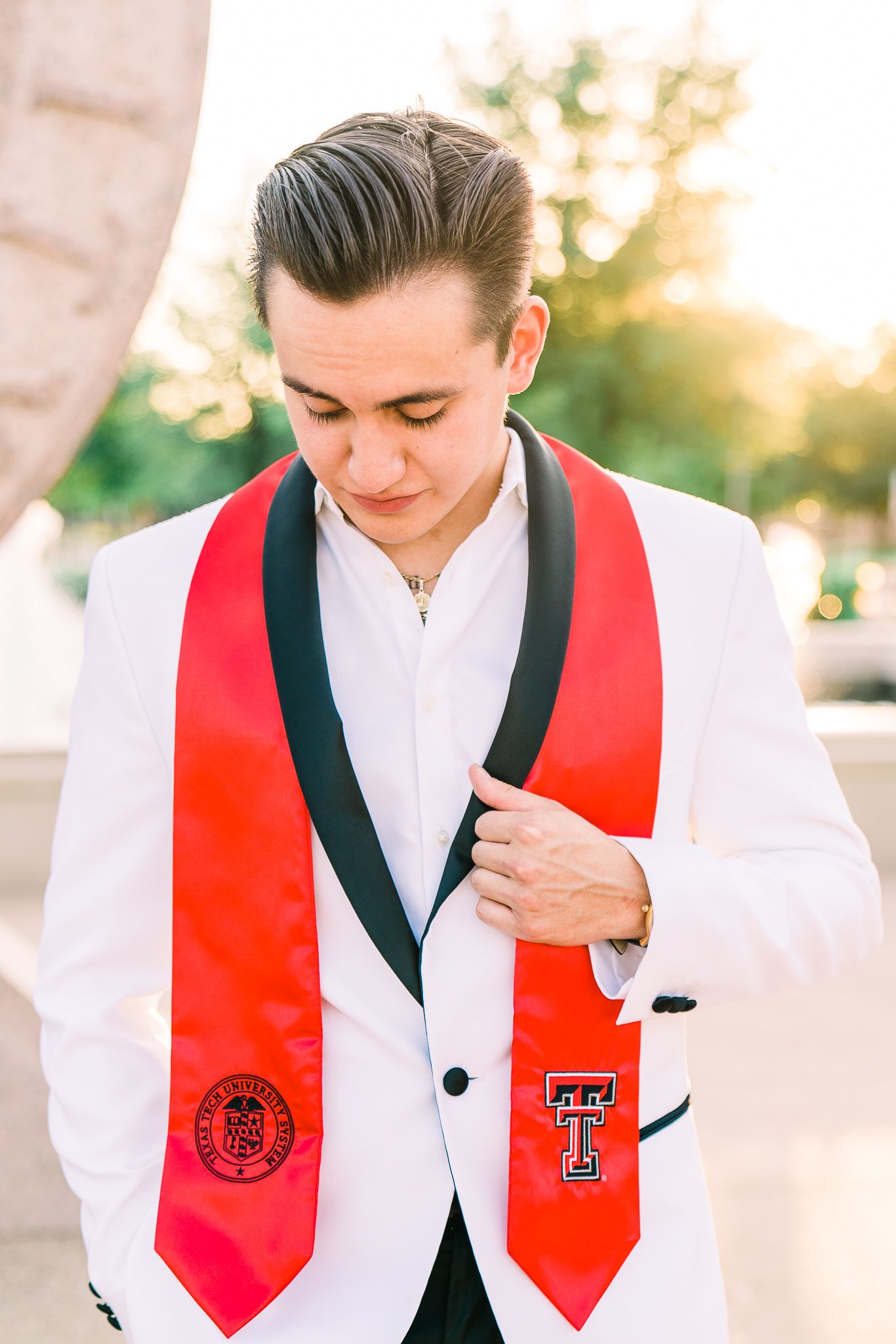 Graduate wearing red and black academic stoles with university emblems adjusts their attire in outdoor setting at sunset.