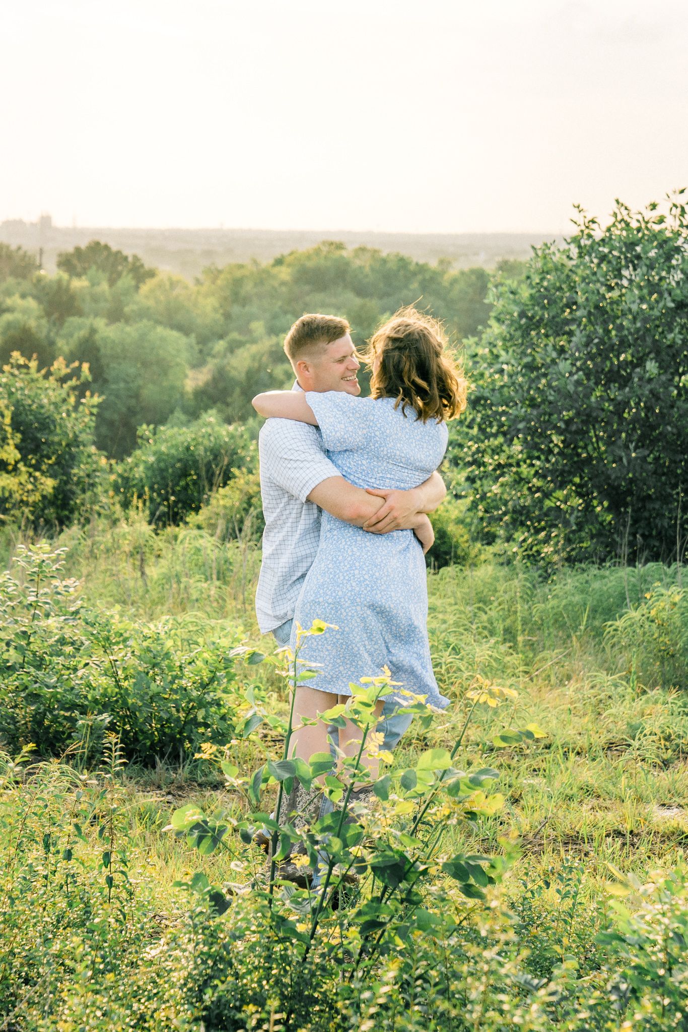 A romantic couple embraces in a sunlit meadow with lush greenery and wildflowers in the background.