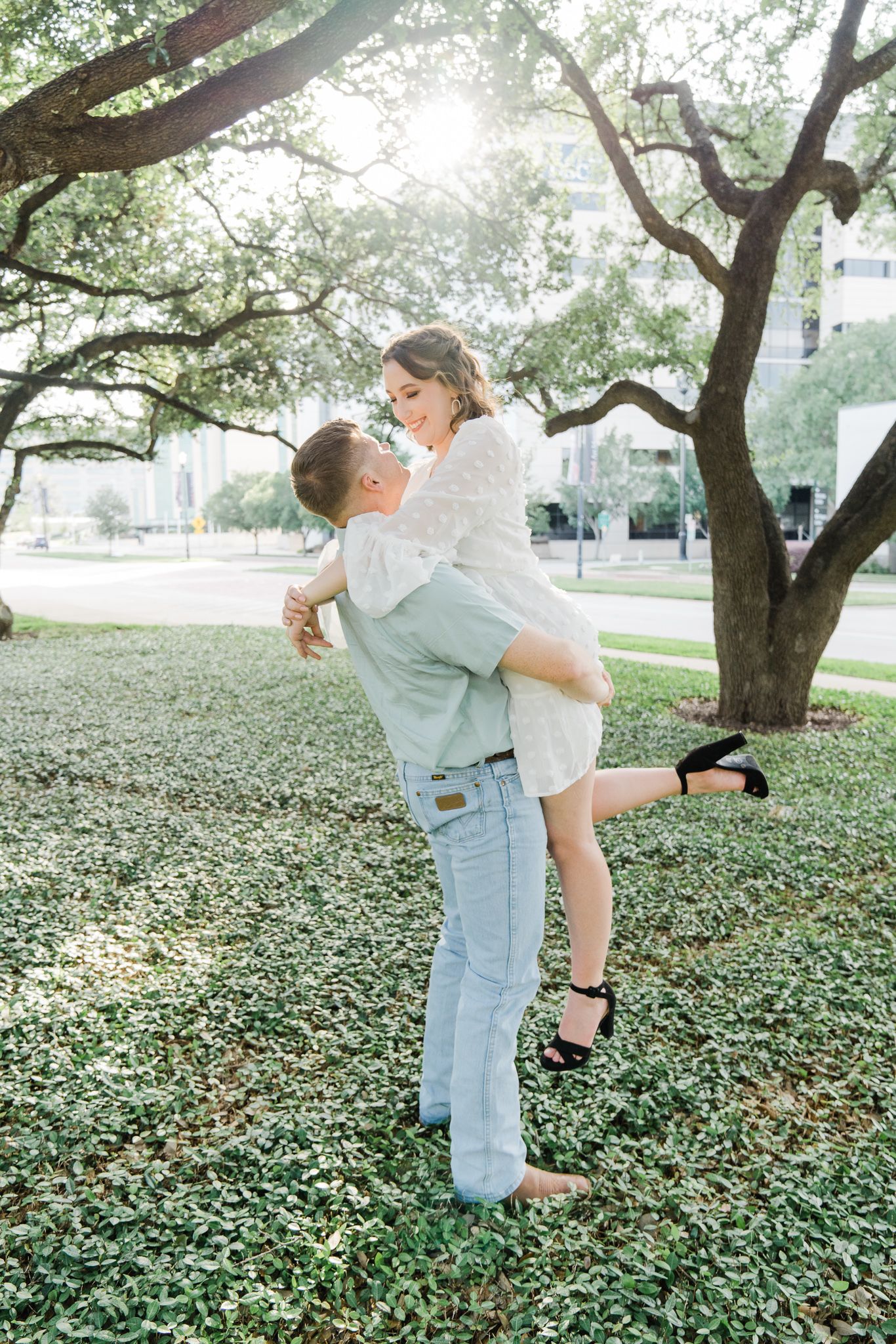 A couple in light blue and white outfits share playful moments under trees in an outdoor photo session.
