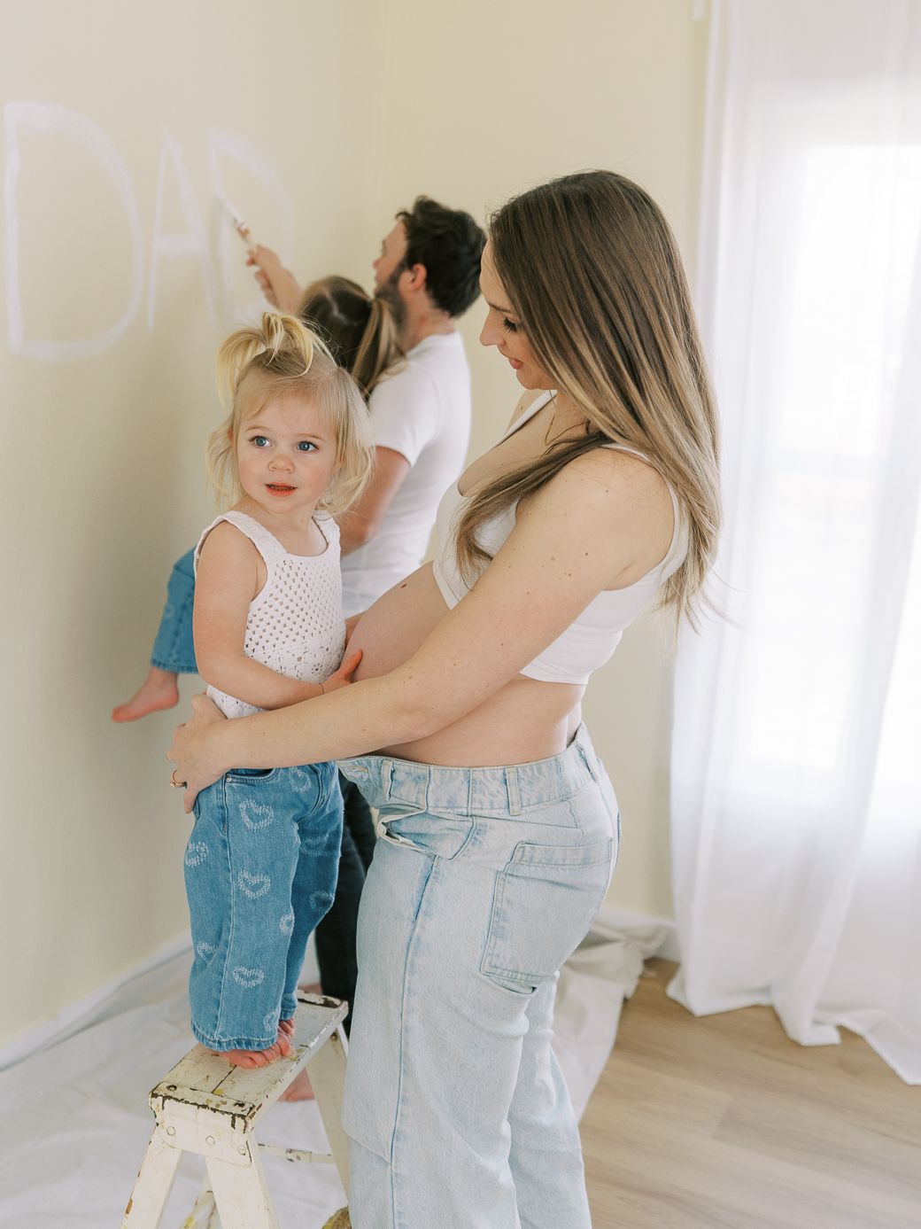 Parent and toddler in denim overalls dance together in a bright white room with natural light streaming in.
