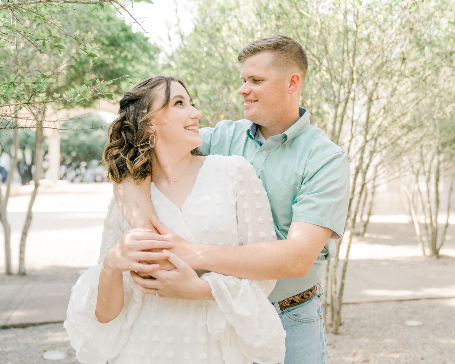 Couple laughing and embracing while dancing outdoors among trees on a bright sunny day.