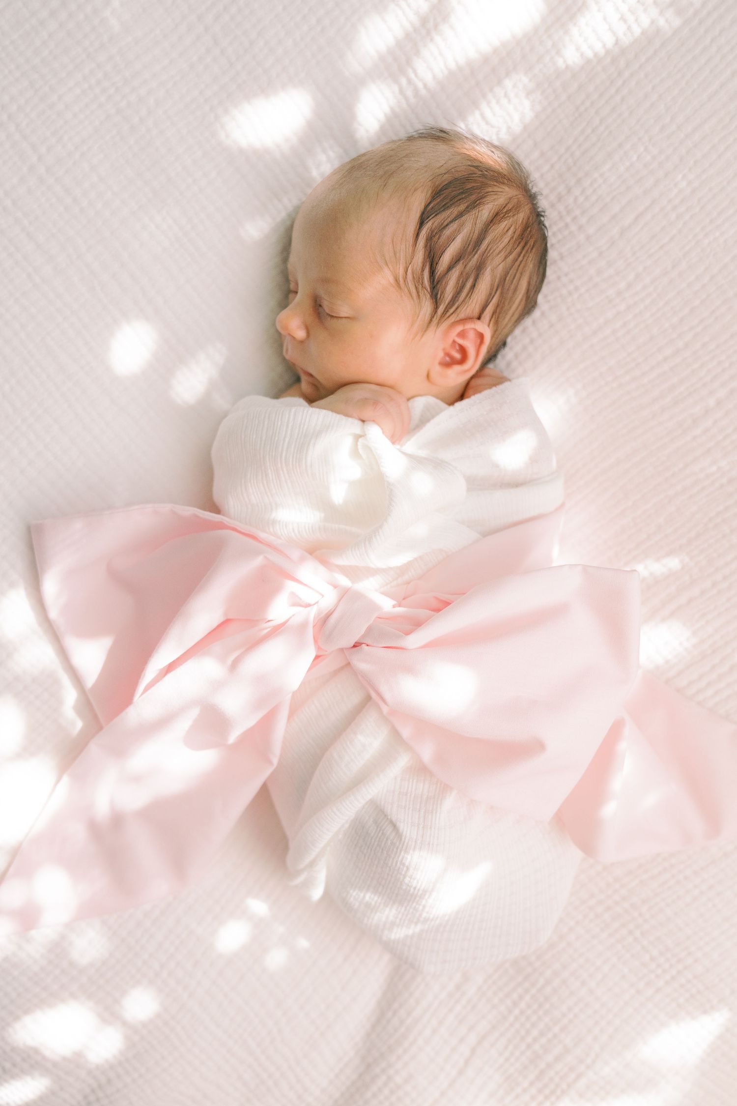 Newborn baby sleeping peacefully on white textured fabric with pink bow details.
