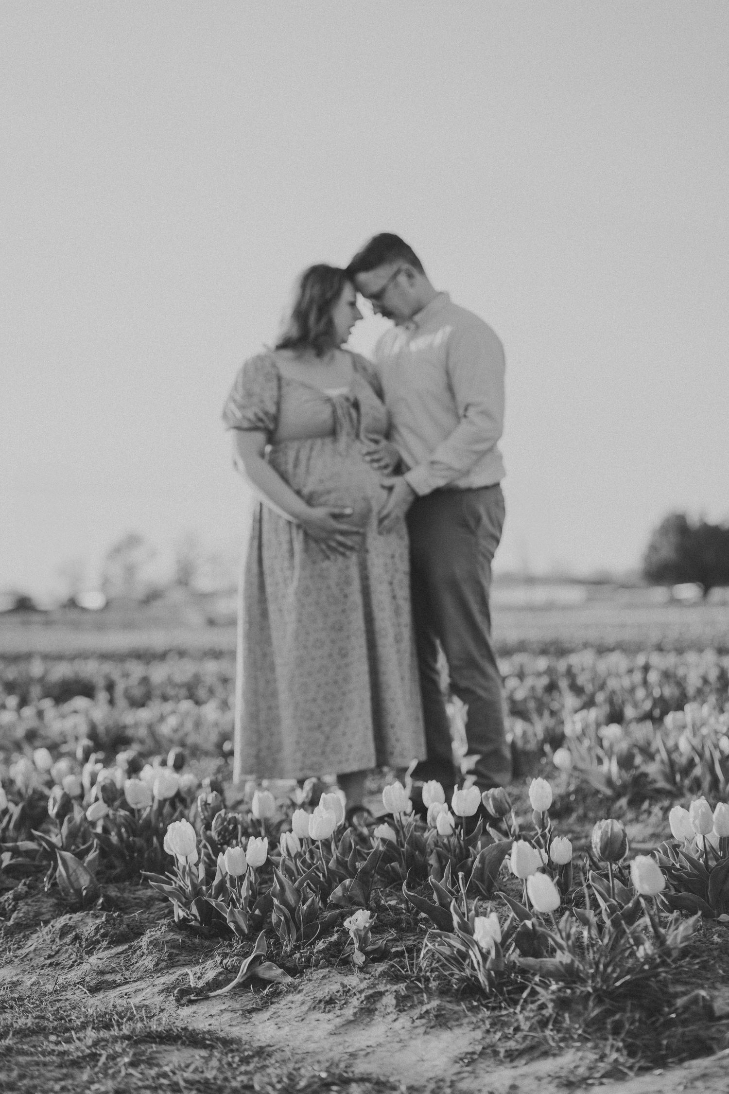 Black and white maternity photo of pregnant couple embracing in a tulip field, touching baby bump tenderly.