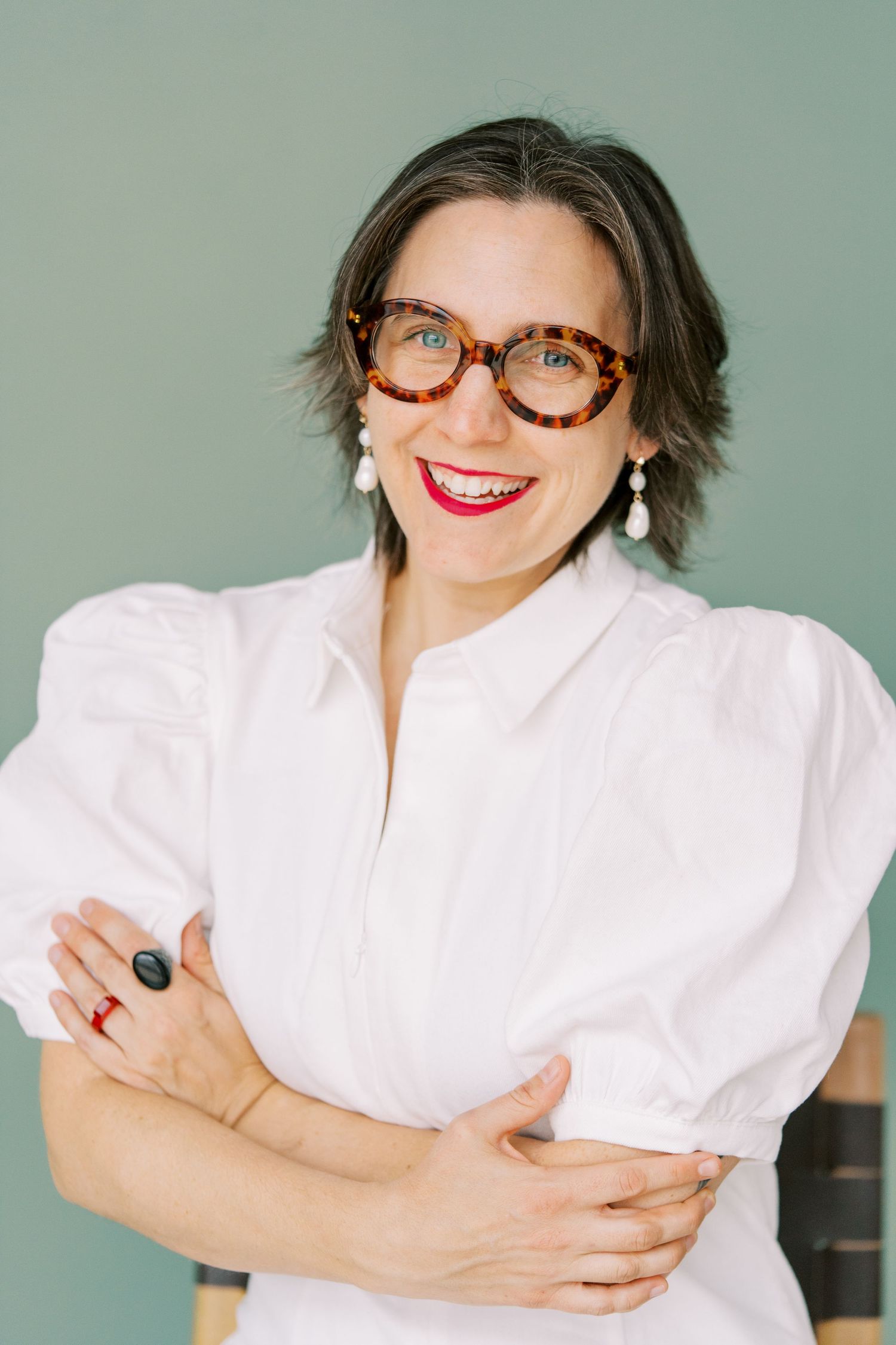 Professional portrait of someone wearing tortoise shell glasses and a white blouse against a sage green background.