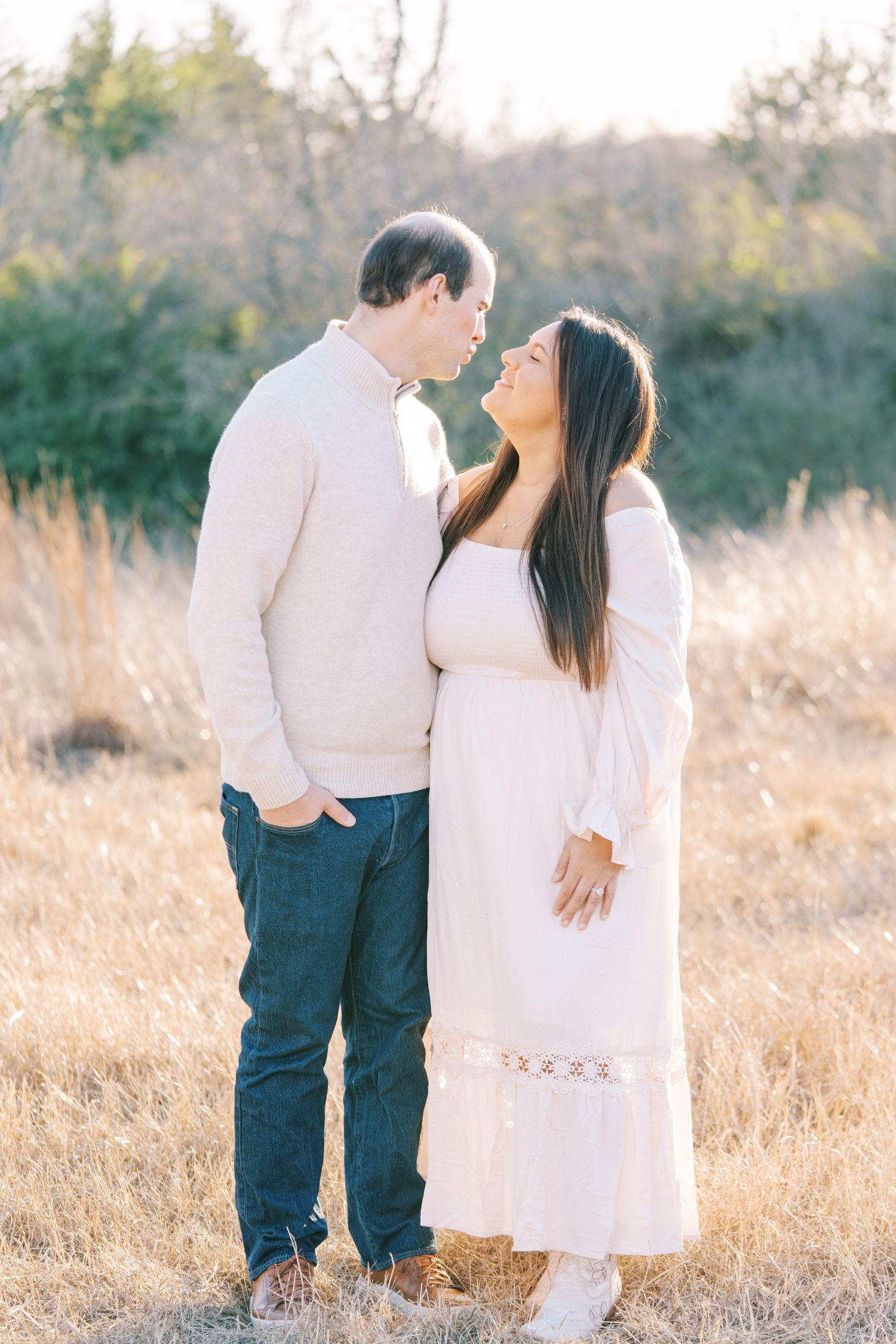 A romantic couple in white outfits share an intimate moment in a sunlit field with trees in background.