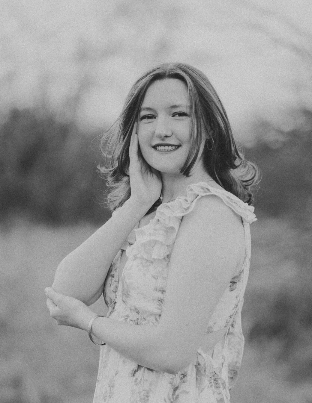 Black and white portrait of someone in a ruffled dress posing with hand near face in natural setting.