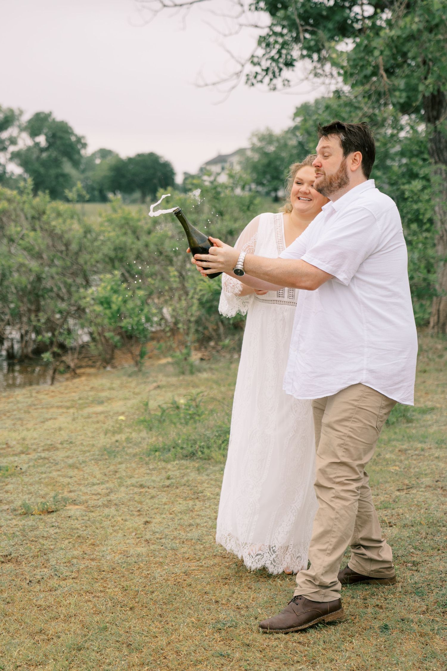 Couple in white celebrating outdoors, popping champagne bottle on lush green lawn with trees in background.