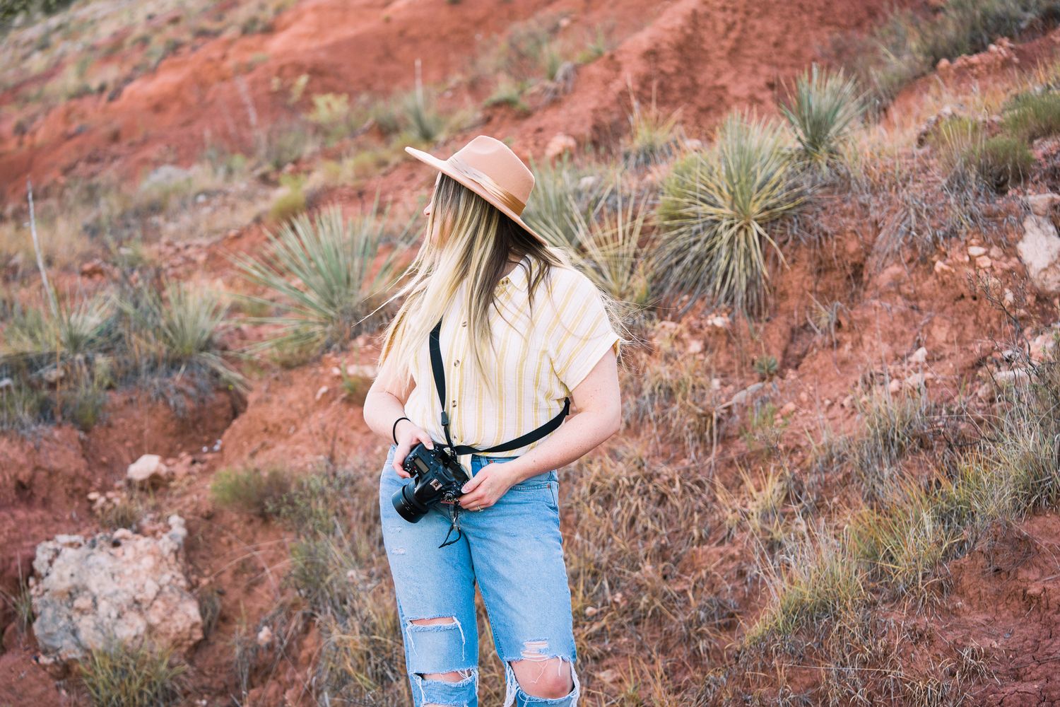A person in blue jeans and a sun hat explores red rock formations in a desert landscape.