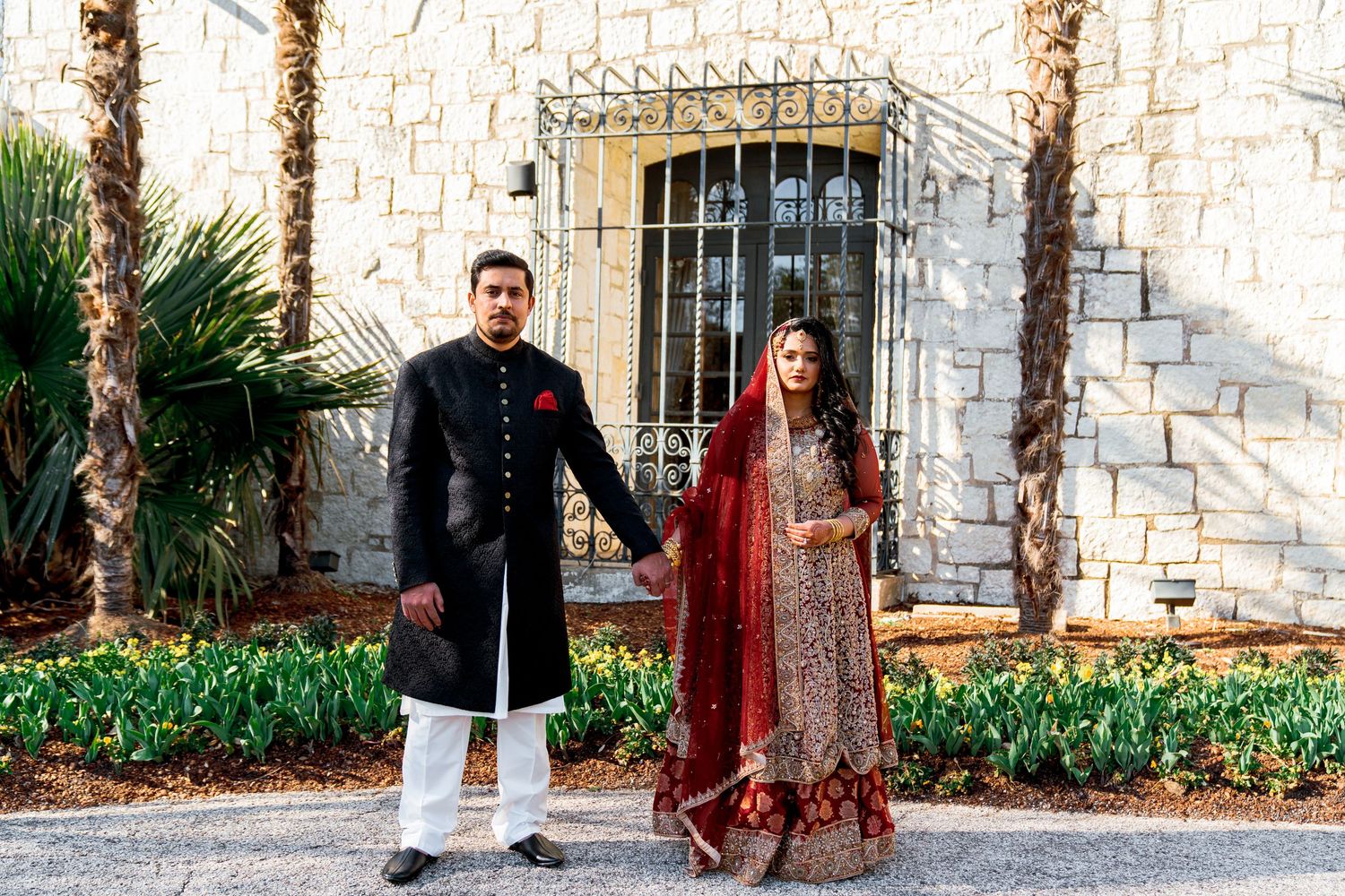 A couple in traditional South Asian wedding attire holds hands in front of a stone wall with palm trees and greenery.