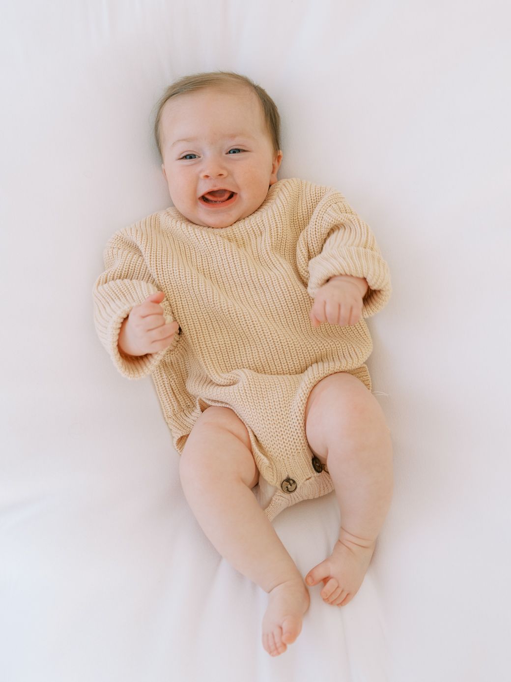 A happy baby wearing a cream colored ribbed romper lies on white fabric smiling at the camera.