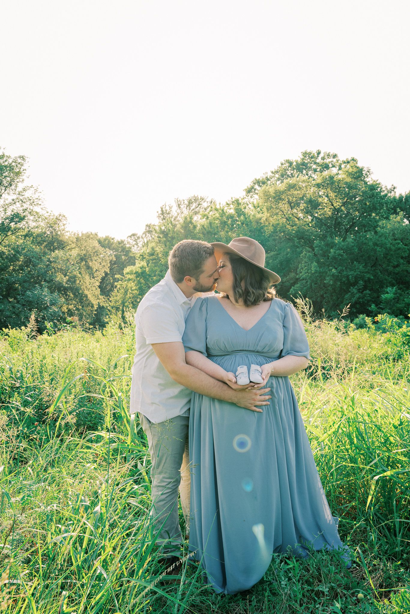 A romantic couple embraces in a sunlit meadow with tall grass and trees, the woman wearing a flowing blue dress and hat.