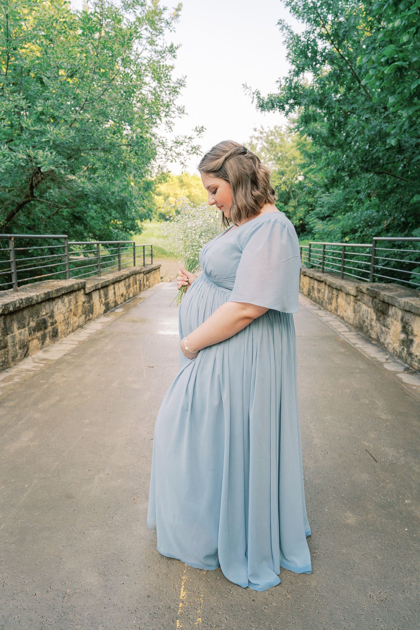 Expectant mother in flowing blue dress poses on stone bridge pathway surrounded by trees.
