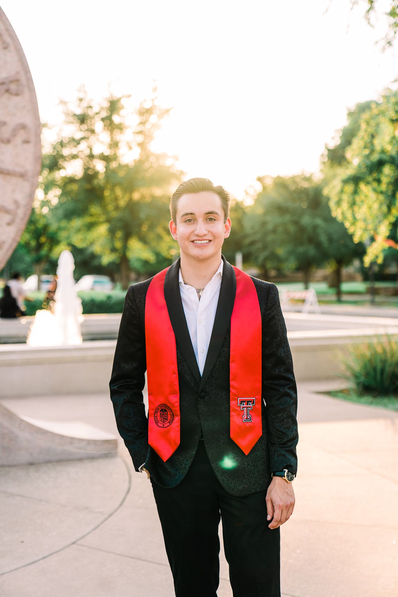 Graduate wearing black suit and red graduation stole poses outdoors during sunset ceremony.