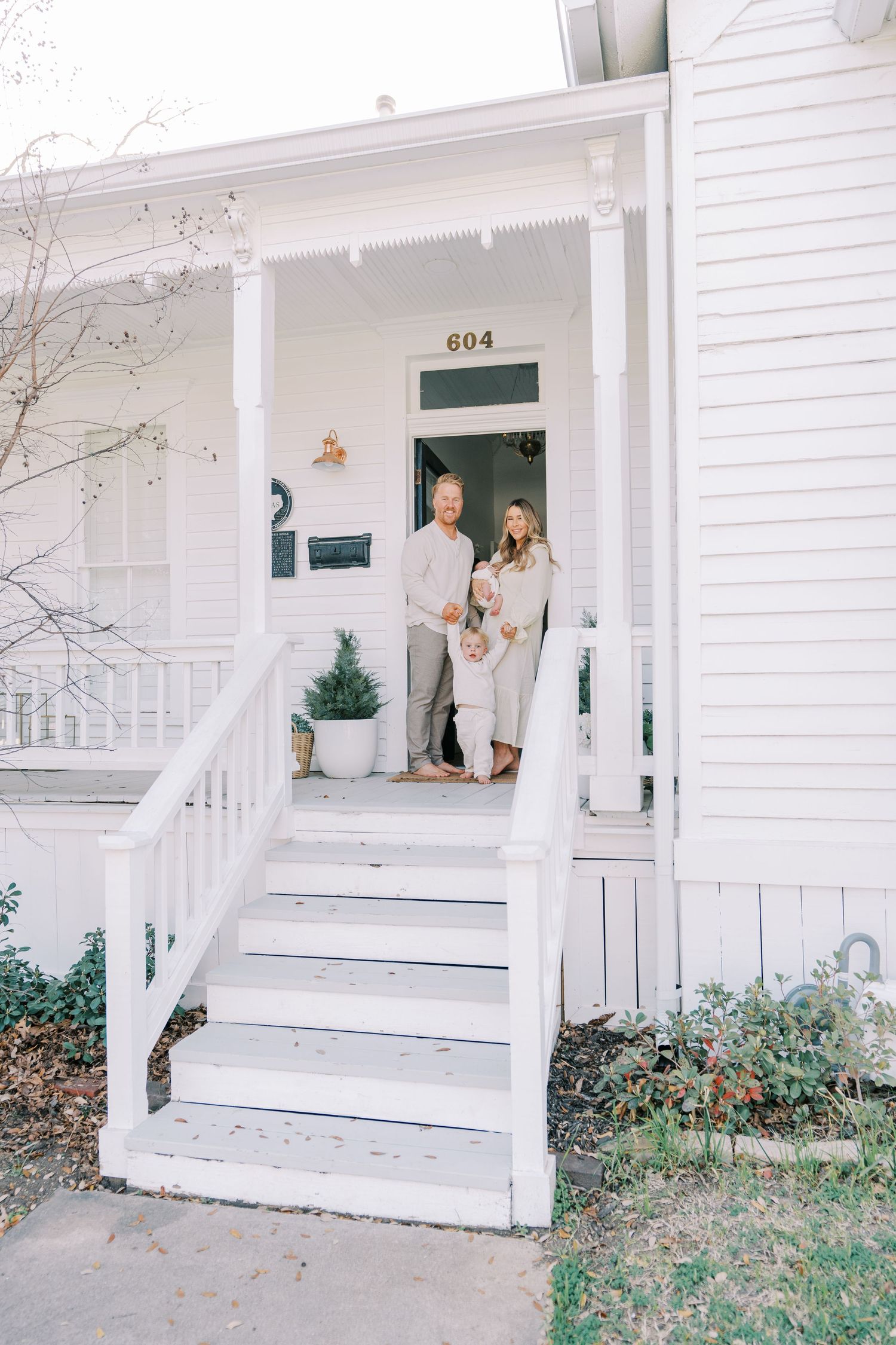 Couple stands on white porch steps of house number 603 in neutral toned clothing.