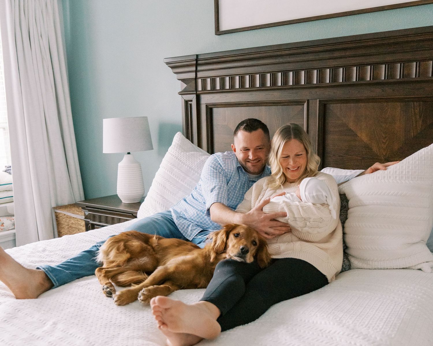 Family relaxing on bed with two dogs while holding newborn baby in cozy bedroom with teal walls.