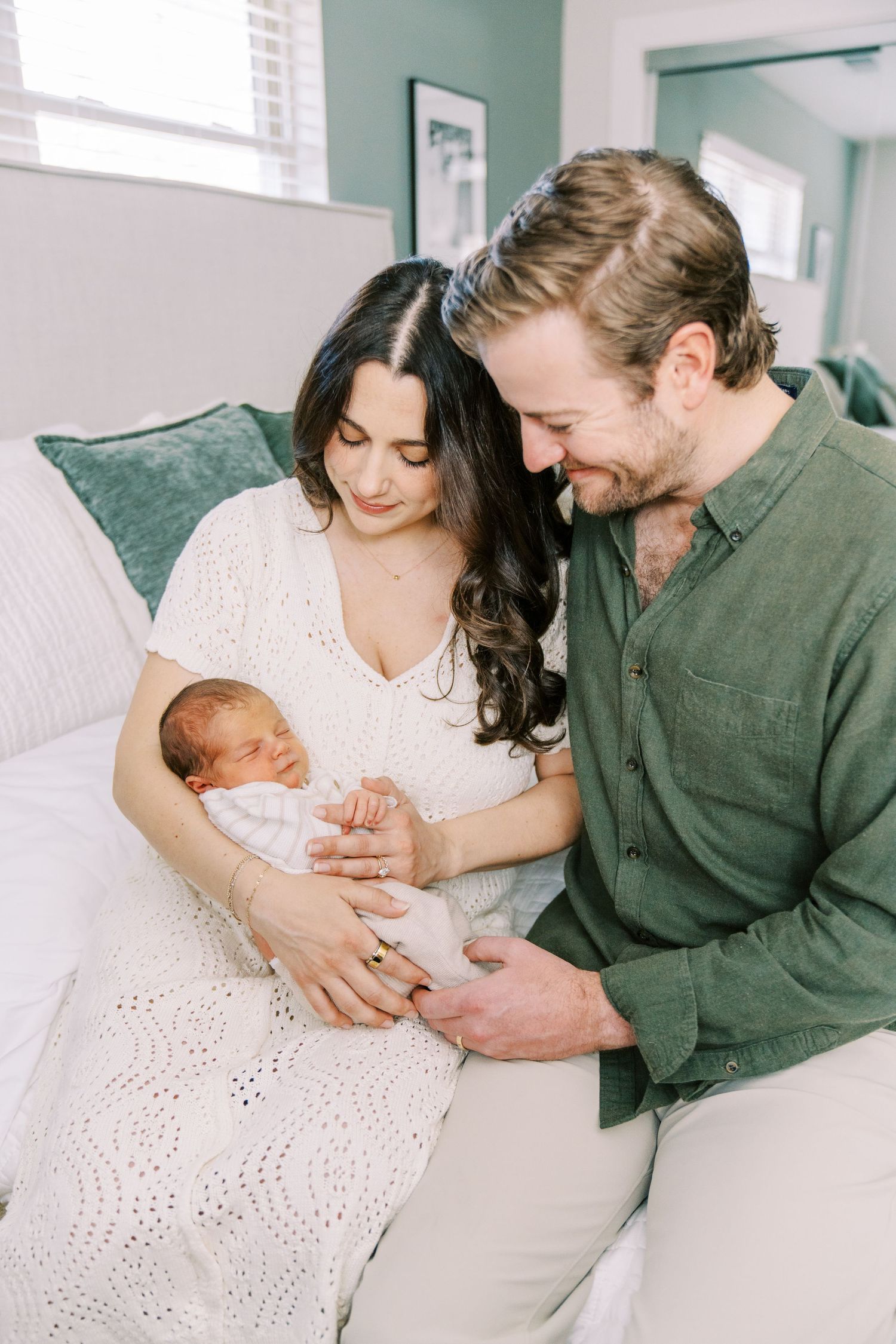 A couple sits together on a white couch admiring their newborn baby in a bright, airy room with mint green walls.
