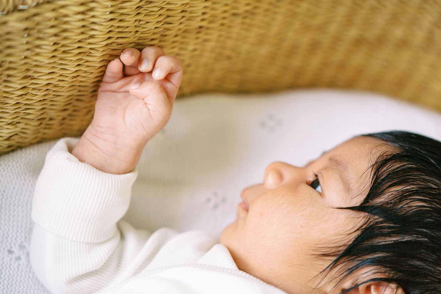 A newborn baby with dark hair lies on its back, reaching up to grasp a woven wicker bassinet in a white onesie.