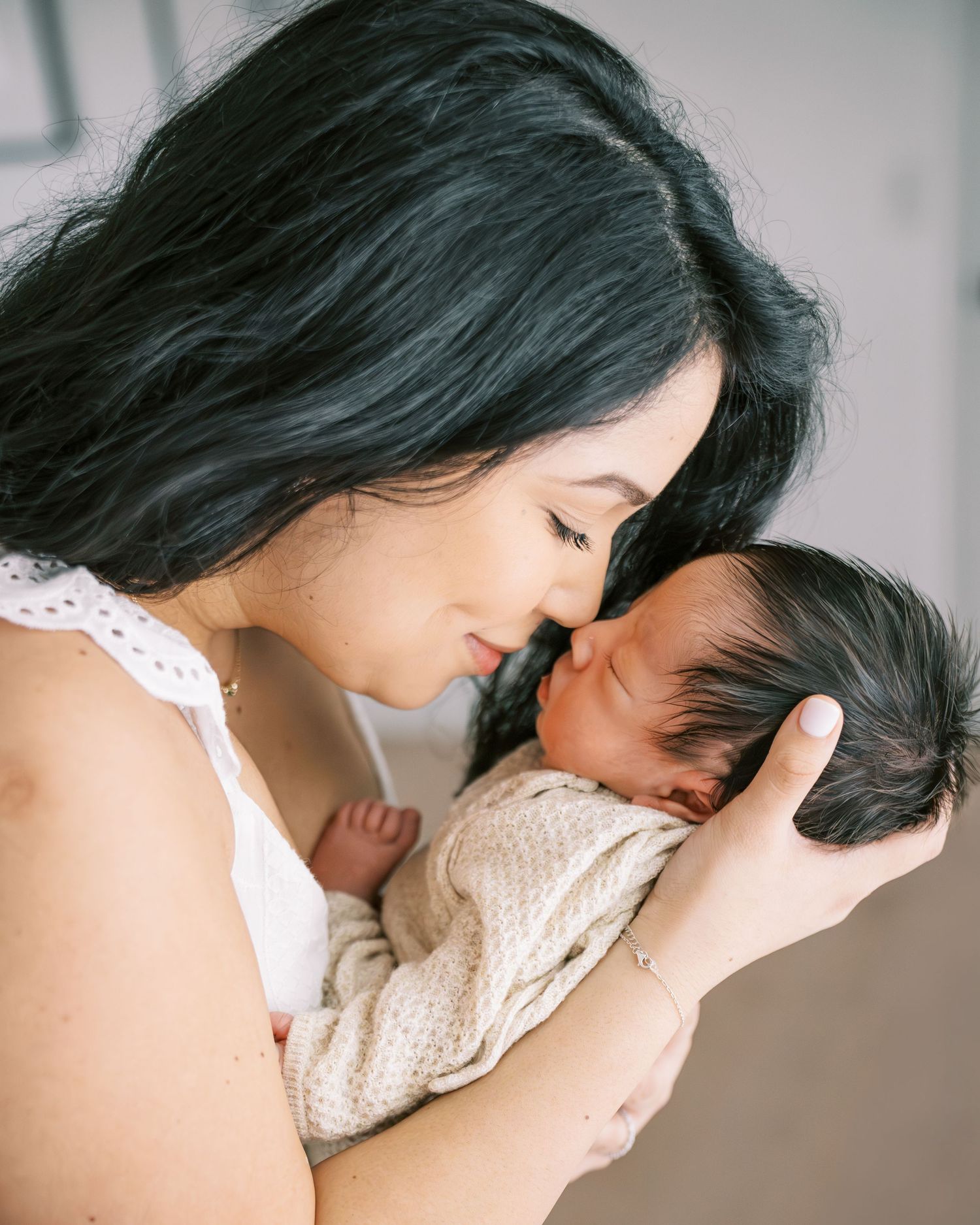 A mother with dark hair tenderly holds and nuzzles her newborn baby wrapped in a knit blanket.