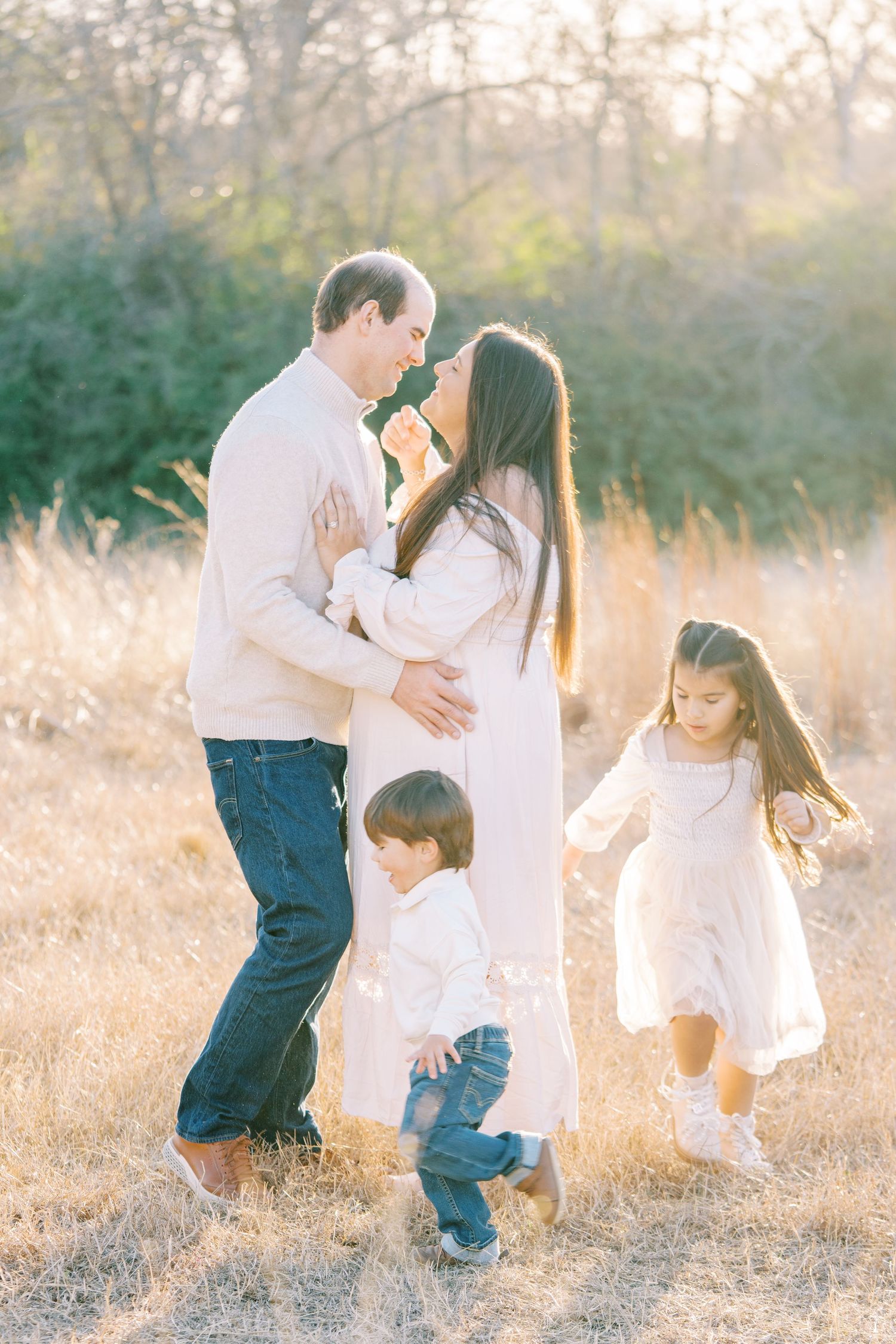 Family of four poses in golden field at sunset, parents sharing a tender moment while children play around them.