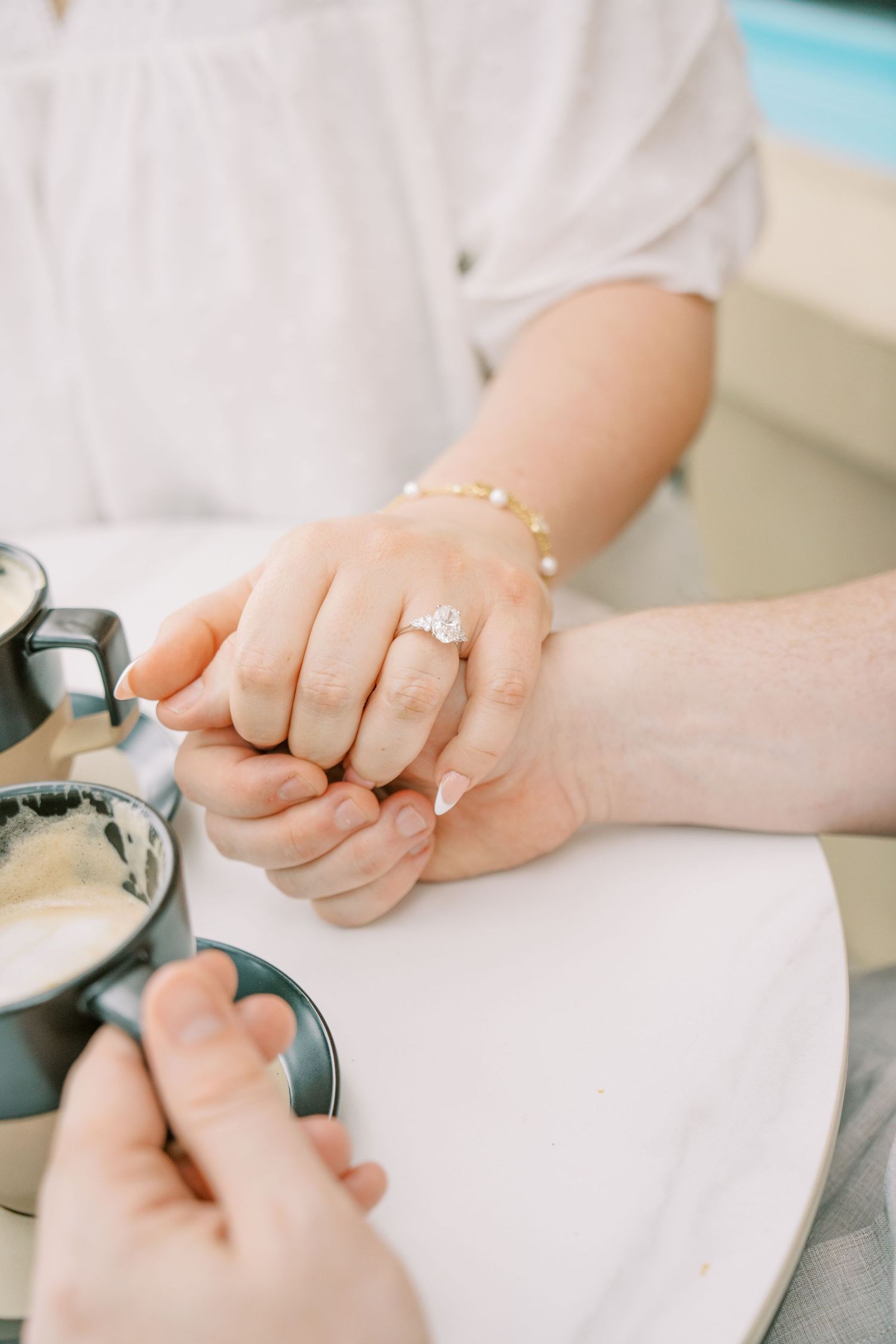 Close-up of womans hand wearing a diamond engagement ring held by her partners hand near coffee cups.