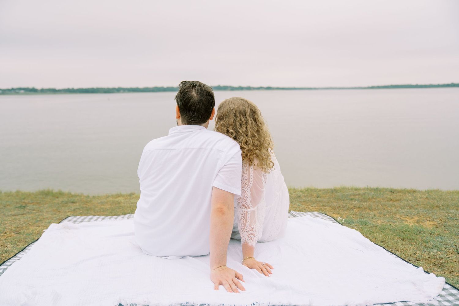 Couple sitting together on a white blanket facing a calm lake, both dressed in white from behind.