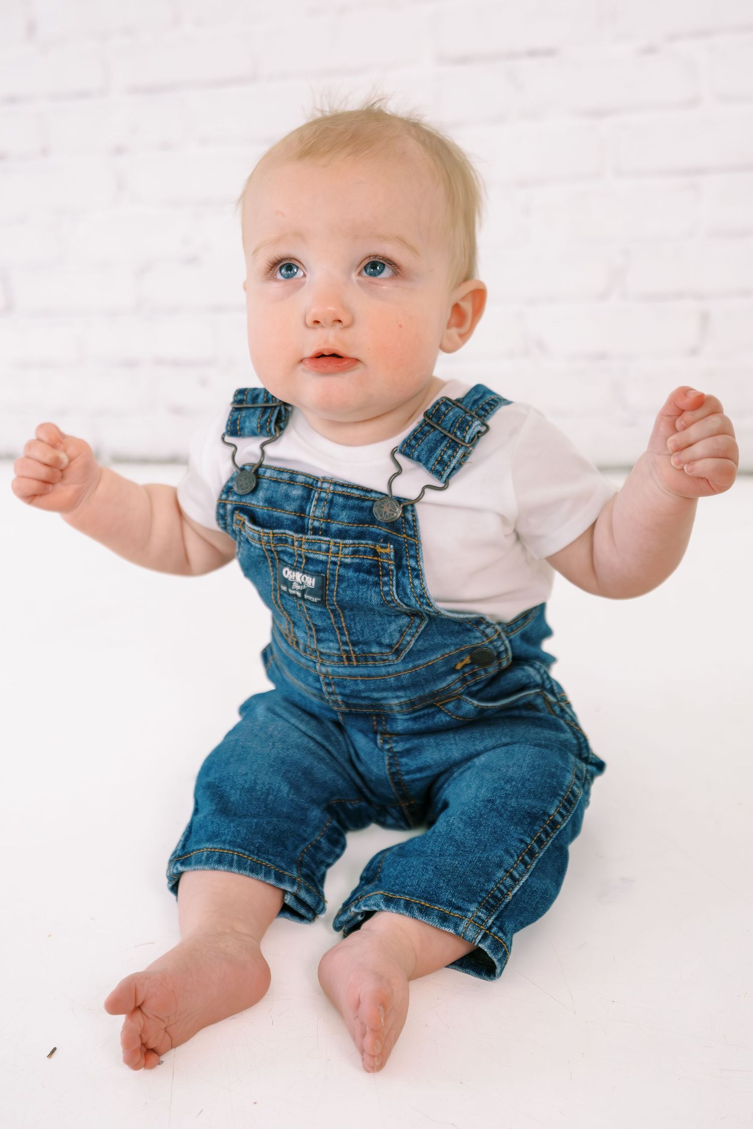 Adorable baby in denim overalls and white shirt sitting barefoot against a white brick wall background.