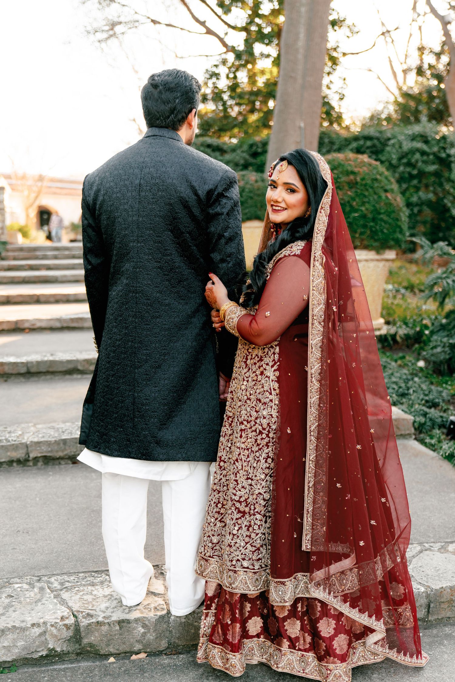 Couple in traditional wedding attire walking together on a stone path during sunset.