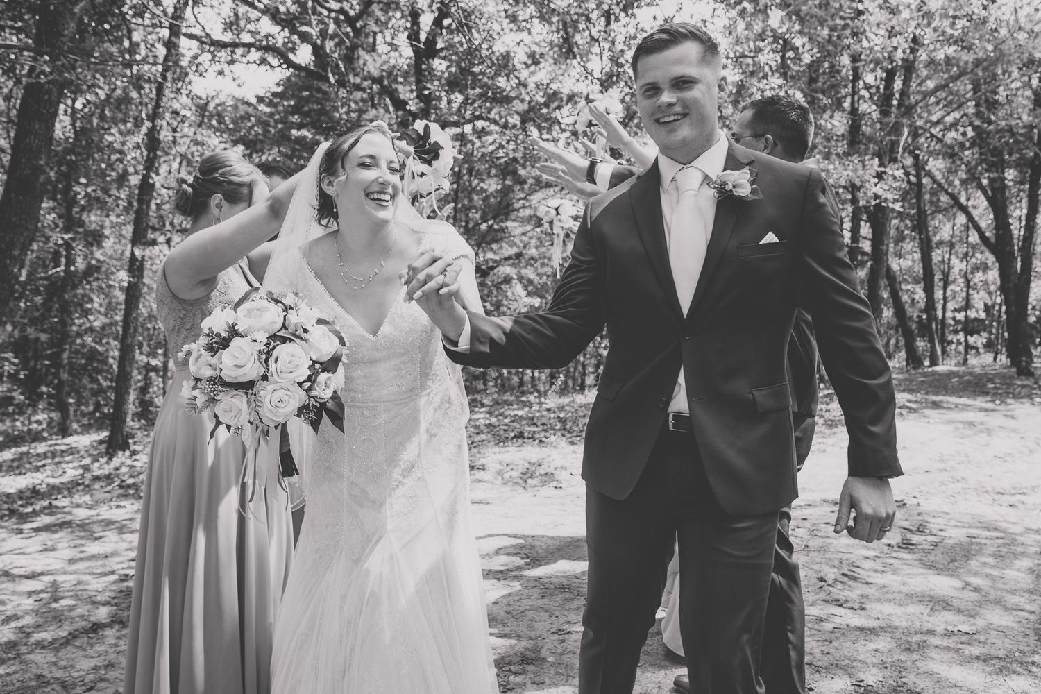 Wedding party walks and laughs together on a wooded path during an outdoor celebration.