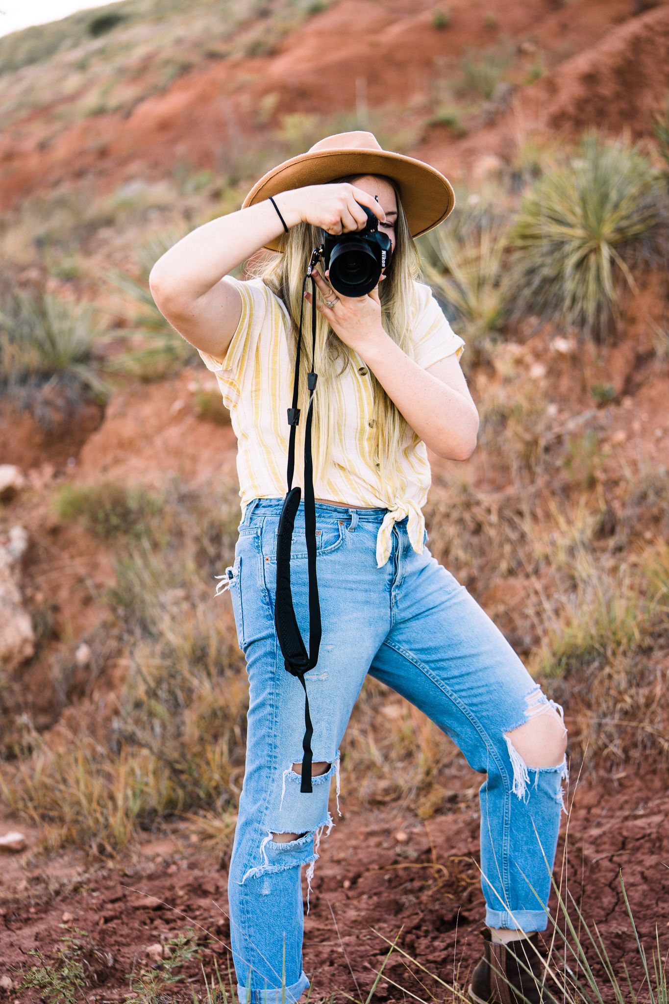 A photographer wearing blue jeans and a straw hat takes photos in a desert landscape with red rocks.