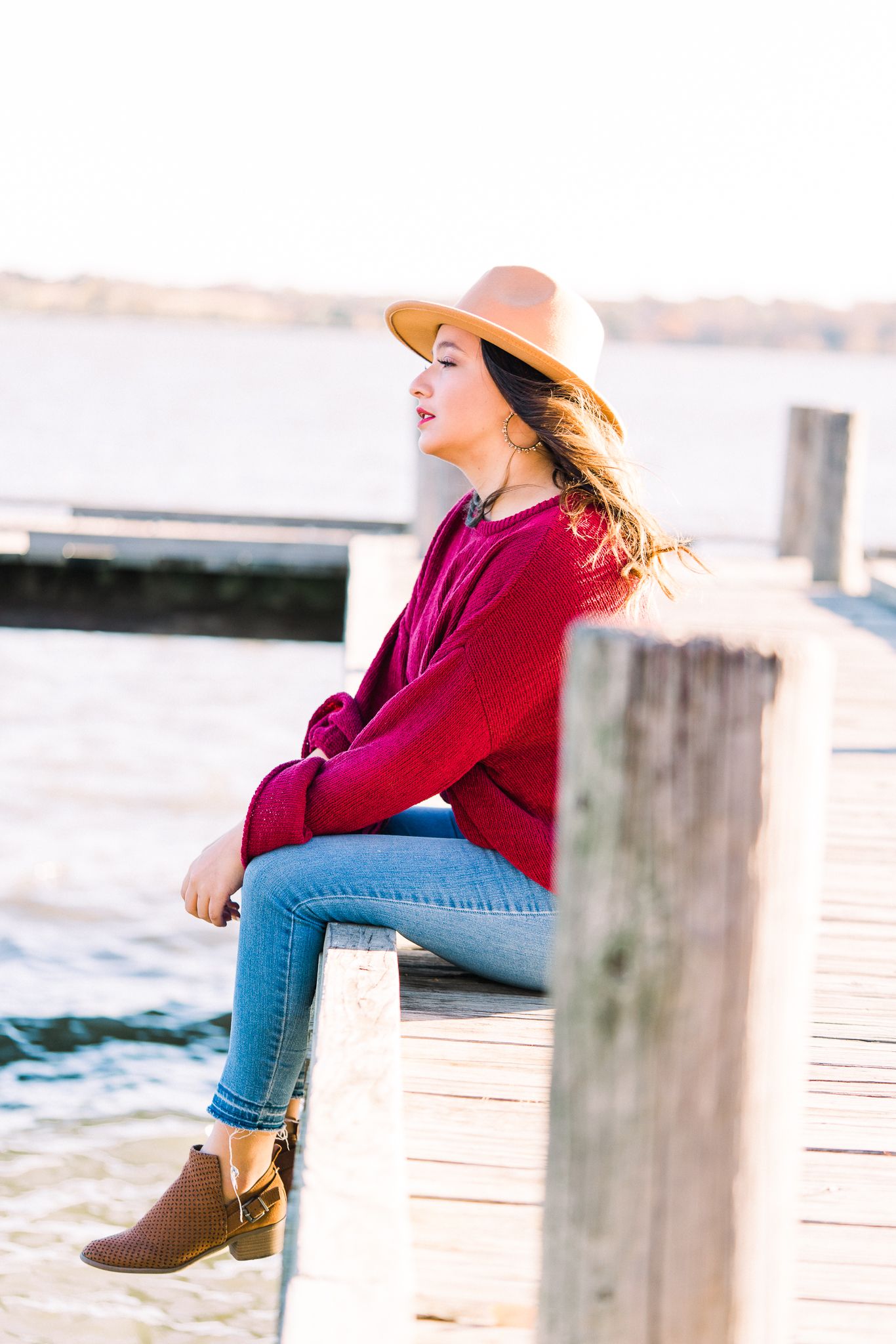 Person in burgundy sweater and jeans sits casually on wooden dock by water at sunset wearing tan wide brim hat.