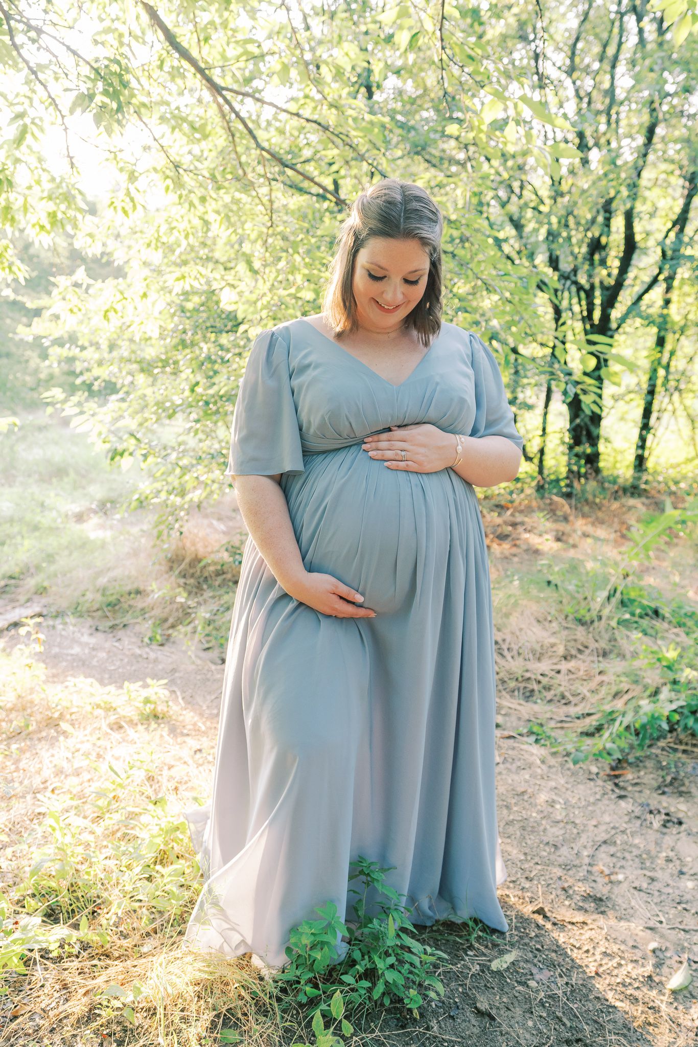 Maternity photoshoot in a dreamy woodland setting with soft sunlight filtering through trees.