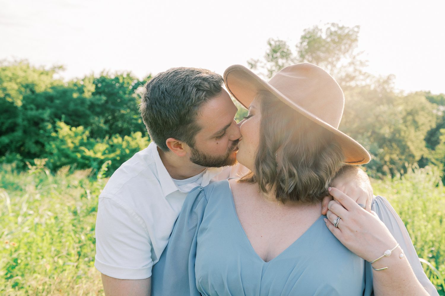 A couple embraces in a sunny meadow with green foliage in the background.